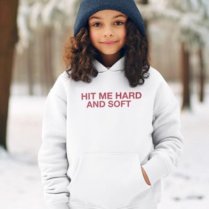 Op de afbeelding: Een persoon draagt een marineblauwe muts en een witte hoodie met de tekst "HIT ME HARD AND SOFT" in bordeauxrood. De hoodie heeft een voorzak en wordt gedragen in een besneeuwde buitenomgeving.
