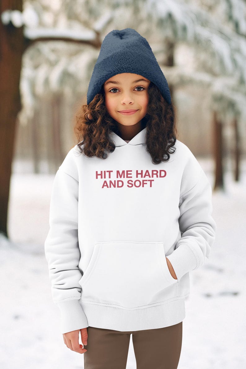 Op de afbeelding: Een persoon draagt een marineblauwe muts en een witte hoodie met de tekst "HIT ME HARD AND SOFT" in bordeauxrood. De hoodie heeft een voorzak en wordt gedragen in een besneeuwde buitenomgeving.