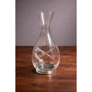 Könnte beinhalten: Eine klare Weindekanter aus Glas mit breitem Boden und schmalem Hals. Der Dekanter hat einen silberfarbenen Boden und ein dekoratives Wirbeldesign im Inneren. Der Dekanter steht auf einer dunklen Holzoberfläche.