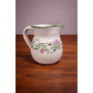 May include: A small, off-white ceramic jug with a handle and a slightly ruffled rim. The jug is decorated with green vines, leaves, and pink flowers. The rim is trimmed with green paint.