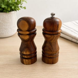 May include: Two matching salt and pepper shakers crafted from dark brown wood with a visible natural grain pattern. Each shaker features a rounded top and a contoured, hourglass-shaped body. The right shaker includes a small silver metal knob.
