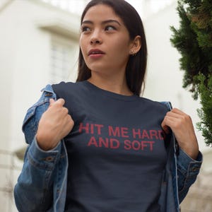 Op de afbeelding: Marineblauw t-shirt met de tekst "HIT ME HARD AND SOFT" in het rood. De persoon draagt een spijkerjasje over het t-shirt. De foto is buiten genomen met een gebouw en groen in de achtergrond.