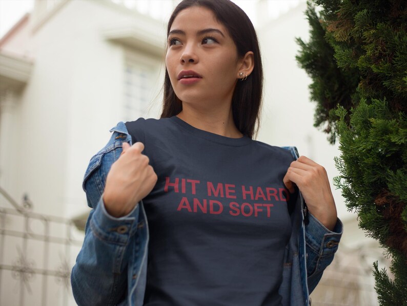 Op de afbeelding: Marineblauw t-shirt met de tekst "HIT ME HARD AND SOFT" in het rood. De persoon draagt een spijkerjasje over het t-shirt. De foto is buiten genomen met een gebouw en groen in de achtergrond.