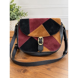 May include: A saddle bag featuring a patchwork design of black, burgundy, and tan suede panels on the front flap. The bag has a black pebbled leather body, a long adjustable shoulder strap, and a central gold-tone metal buckle closure.