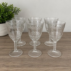 May include: A set of six clear glass goblets featuring a horizontal ribbed pattern on the lower bowl and a decorative laurel leaf band around the upper rim. Each glass has a short, textured stem and a circular base, designed for serving beverages.