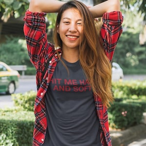 Op de afbeelding: Een donkergrijs t-shirt met de tekst "HIT ME HARD AND SOFT" in het rood. De persoon draagt een rood-zwart geruit flanellen overhemd over het t-shirt en een blauwe spijkerbroek. De persoon glimlacht met de armen achter het hoofd.