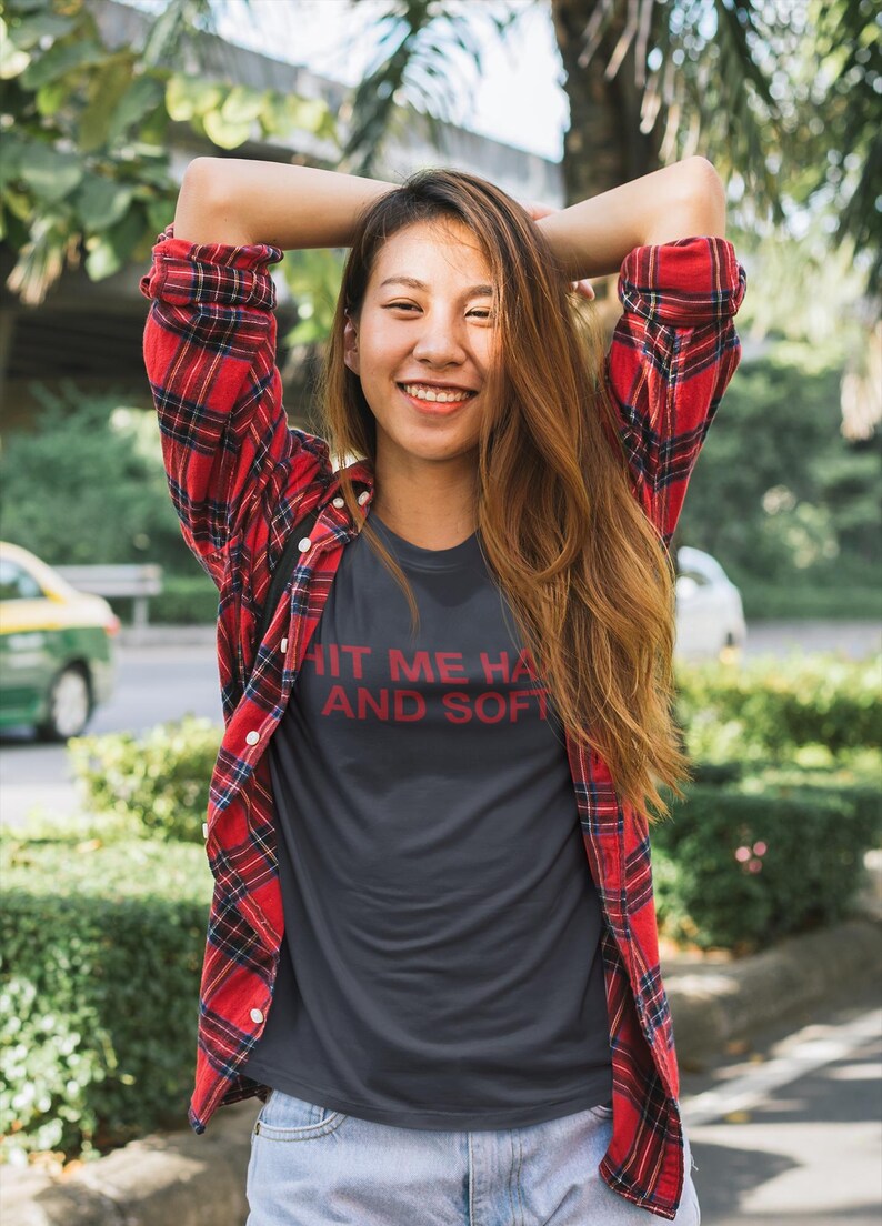 Op de afbeelding: Een donkergrijs t-shirt met de tekst "HIT ME HARD AND SOFT" in het rood. De persoon draagt een rood-zwart geruit flanellen overhemd over het t-shirt en een blauwe spijkerbroek. De persoon glimlacht met de armen achter het hoofd.