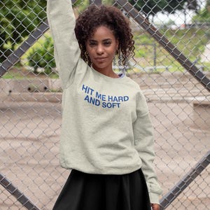Op de afbeelding: Een lichtgrijs sweatshirt met de tekst "HIT ME HARD AND SOFT" in blauw. Het sweatshirt wordt gecombineerd met een zwarte rok. De foto is buiten genomen met een hek van gaas op de achtergrond.