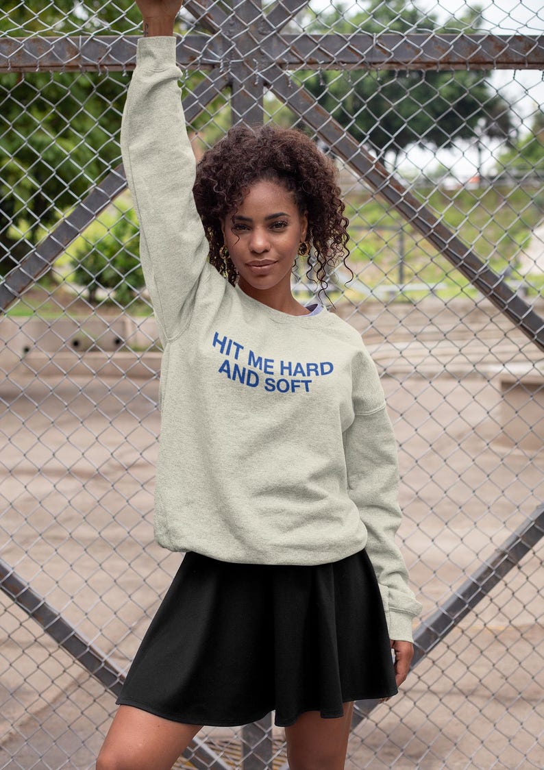 Op de afbeelding: Een lichtgrijs sweatshirt met de tekst "HIT ME HARD AND SOFT" in blauw. Het sweatshirt wordt gecombineerd met een zwarte rok. De foto is buiten genomen met een hek van gaas op de achtergrond.