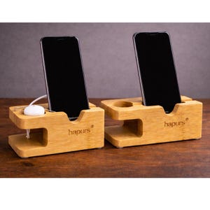 May include: Two bamboo phone stands with a natural wood finish. Each stand holds a black smartphone and features the brand name "hapurs". One stand has a charging dock for a smartwatch or earbuds.