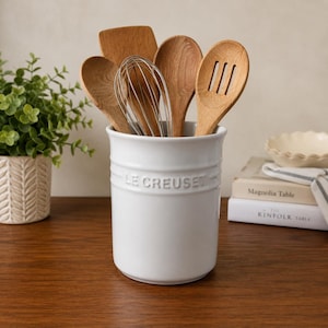 Puede incluir: Un bote para utensilios de cerámica blanca Le Creuset lleno de varios utensilios de cocina de madera, incluyendo una espátula, dos cucharas, una cuchara ranurada y un batidor de alambre metálico. El recipiente cilíndrico lleva el nombre de la marca.