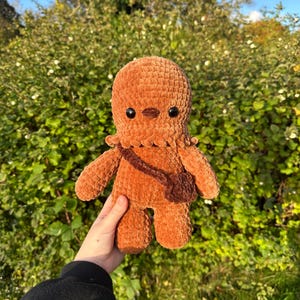 Op de afbeelding: Een handgemaakte, bruine gehaakte pop met zwarte ogen en een kleine bruine tas. Het pluche speelgoed heeft een getextureerd, fuzzy uiterlijk en wordt tegen een achtergrond van groen gebladerte gehouden.