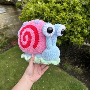 May include: A crocheted snail toy, resembling Gary from Spongebob Squarepants. The snail has a pink shell with a red spiral, a blue body, and large eyes. The toy is held against a backdrop of green grass and foliage.