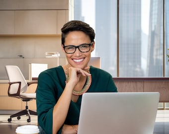 May include: A woman wearing glasses and a green shirt smiles while sitting at a desk with a laptop and a cup of coffee. She is in a modern office setting with a window behind her.