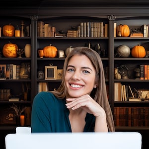 May include: A woman with long brown hair smiles while sitting in front of a dark wooden bookshelf decorated with pumpkins and books.