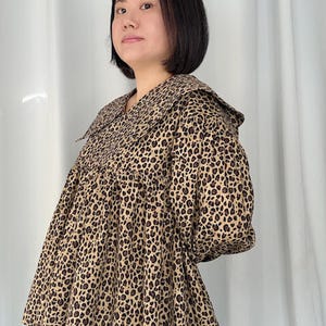 May include: A tan and brown leopard print top with a large collar. The top has a loose, flowing fit and short sleeves. The fabric has a repeating pattern of dark brown spots on a tan background. The collar is wide and flat, and the top appears to be made of a soft material.
