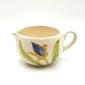 May include: Cream-colored ceramic creamer with a handle and spout. The creamer is decorated with a blue and yellow floral design and green leaves. The creamer is on a white background.