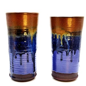 May include: Two handmade ceramic tumblers. The top of each tumbler is a metallic bronze color, transitioning to a deep blue with dripped glaze effects. The base is a dark brown. The tumblers are cylindrical and stand upright.