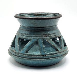 May include: A teal ceramic candle holder with a geometric design. The holder has a wide base, a cylindrical body with cut-out shapes, and a flared top. The rim is dark brown. The piece is on a white background.