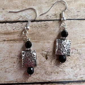 May include: A pair of silver earrings with black beads and a square charm with a floral design.