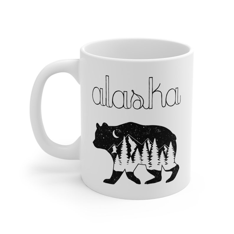 This custom-made design is printed on both sides of an 11oz ceramic mug for coffee or tea, and features the word Alaska in vibey black cursive font, and a grizzly bear adorned with mountains and evergreen trees under a crescent moon.
