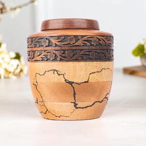 May include: A wooden urn with a dark brown lid and a decorative floral band. The body of the urn is made of light and medium-toned wood with dark veining. The urn is designed to hold ashes.