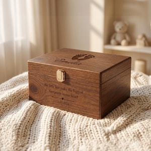 May include: A rectangular wooden keepsake box with a gold-tone metal latch. The lid features engraved baby footprints and the name Emelia. The front is engraved with the text: the little feet make the biggest footprints in our heart 21.05.2024.