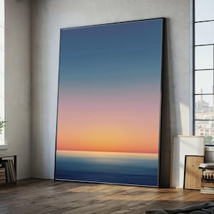 May include: A large framed art print showing a seascape. The artwork displays a gradient of colours, shifting from a deep blue at the bottom to a vibrant orange and pink at the horizon, with a clear blue sky above. The frame is black.