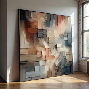 Könnte beinhalten: Großformatiges, gerahmtes abstraktes Gemälde. Das Kunstwerk zeigt ein strukturiertes, geschichtetes Design mit Pinselstrichen in Beige-, Grau-, Braun- und Blautönen. Es lehnt an einer weißen Wand in einem Raum mit Holzboden und Fenster.