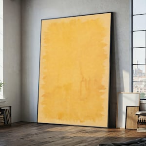 May include: Large framed abstract art print. The artwork features a textured, yellow-orange background with a black frame. The piece is leaning against a wall in a room with a window and wooden floor.