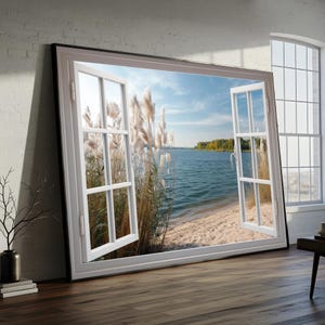 Könnte beinhalten: Großformatige gerahmte Kunst, die ein offenes Fenster mit Blick auf eine ruhige See zeigt. Der weiße Fensterrahmen zeigt hohe Schilfgräser, einen Sandstrand und blaues Wasser unter einem hellen Himmel. Das Kunstwerk steht auf einem Holzboden.