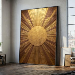 May include: Large, framed wall art featuring a sunburst design. The artwork has a central gold circle with radiating lines in alternating gold and brown tones. The frame is black, and the piece is leaning against a wall on a wooden floor.