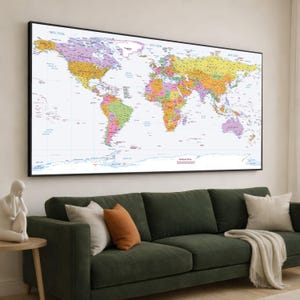 Op de afbeelding: Een grote, ingelijste wereldkaart met een zwarte rand, waarop landen in verschillende kleuren worden weergegeven. De kaart hangt boven een groene bank met kussens en een deken. Links staat een kleine tafel met een beeldhouwwerk.