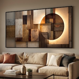 Op de afbeelding: Abstracte wandkunst met een geometrisch ontwerp en een grote, verlichte cirkel. Het kunstwerk is samengesteld uit verschillende getextureerde materialen in tinten bruin, beige en goud, omlijst in zwart. Het hangt boven een beige bank met decoratieve kussens.