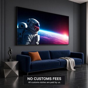 Op de afbeelding: Een rechthoekige wandkunstprint met een astronaut in een wit pak die uitkijkt over een gloeiende planeet met een levendige rode en blauwe horizon tegen een donkere sterrenhemel, ingelijst in een dunne zwarte lijst boven een marineblauwe bank.