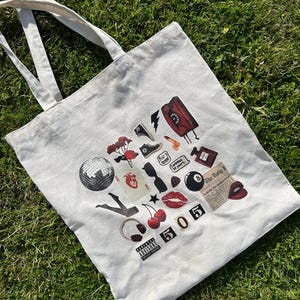 Könnte beinhalten: Eine weiße Canvas-Tragetasche mit einem rot-schwarz-weißen Design. Das Design beinhaltet eine Discokugel, Kirschen, ein rotes Herz, ein rotes Telefon, ein Paar schwarze Converse-Schuhe, eine schwarze 8-Ball, roten Lippenstift und den Text "505".