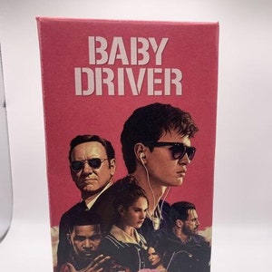 May include: A red DVD case with the title "BABY DRIVER" in white block letters. The case features a collage of actors, including a man wearing sunglasses and earbuds. The overall theme is a movie.