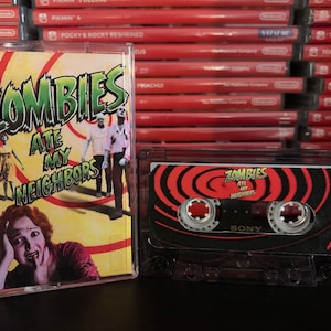 May include: A clear cassette tape and case featuring the title "Zombies Ate My Neighbours" in green and red. The case shows a woman with a terrified expression and zombies. The cassette tape has a red and black design.