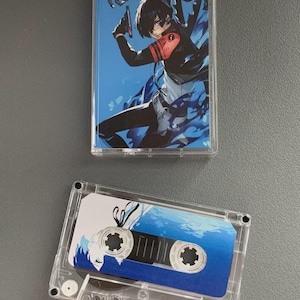 Puede incluir: Dos cintas de casete transparentes. Una cinta presenta una ilustración azul y blanca de un personaje sosteniendo un arma. La otra cinta tiene un diseño azul y blanco. Ambas cintas están sobre una superficie gris.
