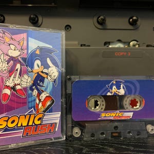 Puede incluir: Dos cintas de cassette con personajes de Sonic the Hedgehog. Una cinta tiene una portada morada y azul con Sonic y Blaze, y el texto "SONIC RUSH". La otra cinta tiene un diseño azul y morado con Sonic y el texto "SONIC RUSH".