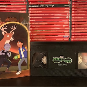 May include: An "Infinity Train Book Two" VHS tape and a matching book cover featuring cartoon characters and a deer. The tape is black with a green and white logo. A stack of red Nintendo Switch game cases is in the background.