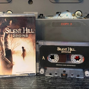 May include: A clear cassette tape of the "Silent Hill Origins" original game soundtrack. The cover art features a man with a weapon and a figure in a foggy landscape. The cassette case is next to the tape, with the text "COPY 3" visible.