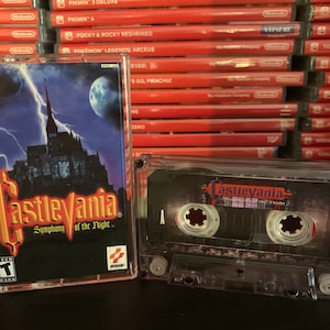 May include: A clear cassette tape of the video game Castlevania: Symphony of the Night, with a cover featuring a castle, lightning, and a moon. The cassette is in a clear case, with the game title visible. A stack of Nintendo Switch game cases is in the background.