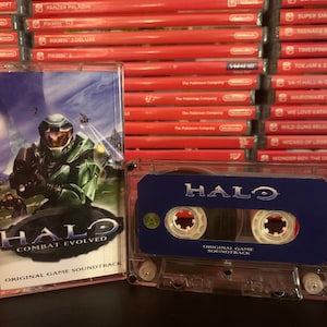 May include: A clear cassette tape case with a blue and white label that reads "HALO" and "ORIGINAL GAME SOUNDTRACK". The cover art features a green armored figure with the text "HALO COMBAT EVOLVED". A stack of red video game cases is in the background.