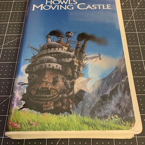 Puede incluir: Una cinta VHS de la película de animación "El castillo ambulante" de Hayao Miyazaki. La portada presenta una ilustración detallada del castillo con chimeneas sobre un fondo de cielo azul y montañas. La cinta está en una caja blanca.