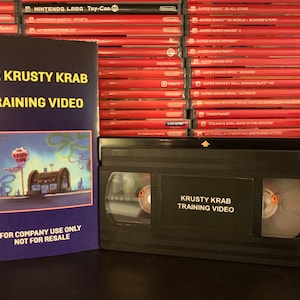 May include: A black VHS tape and a matching sleeve with the text "THE KRUSTY KRAB TRAINING VIDEO". The sleeve has an image of the Krusty Krab restaurant. The text "FOR COMPANY USE ONLY NOT FOR RESALE" is also visible. A stack of red video game cases is in the background.