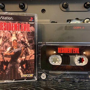 May include: A clear plastic case containing a PlayStation game, "Resident Evil," with a man holding a gun on the cover. Next to it is a cassette tape with the game title in red. The background includes a stereo.