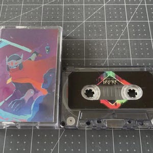 Puede incluir: Una cinta de cassette transparente y su estuche. El estuche presenta una colorida ilustración de una figura con una espada. La cinta de cassette tiene la palabra "DRIFTER" en un diseño colorido.