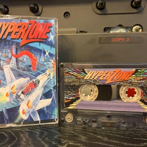 Könnte beinhalten: Zwei Vintage-Kassetten. Eine hat ein farbenfrohes Cover mit dem Wort "HYPERZONE" in Rot und Orange. Die andere Kassette hat ein lila Etikett mit demselben Titel. Die Kassetten befinden sich in transparenten Kunststoffhüllen.