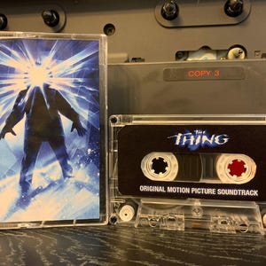 May include: A clear cassette tape case with a movie poster design featuring a silhouetted figure with a bright light emanating from the head. The cassette tape next to it has the movie title "The Thing" and "Original Motion Picture Soundtrack" printed on it.
